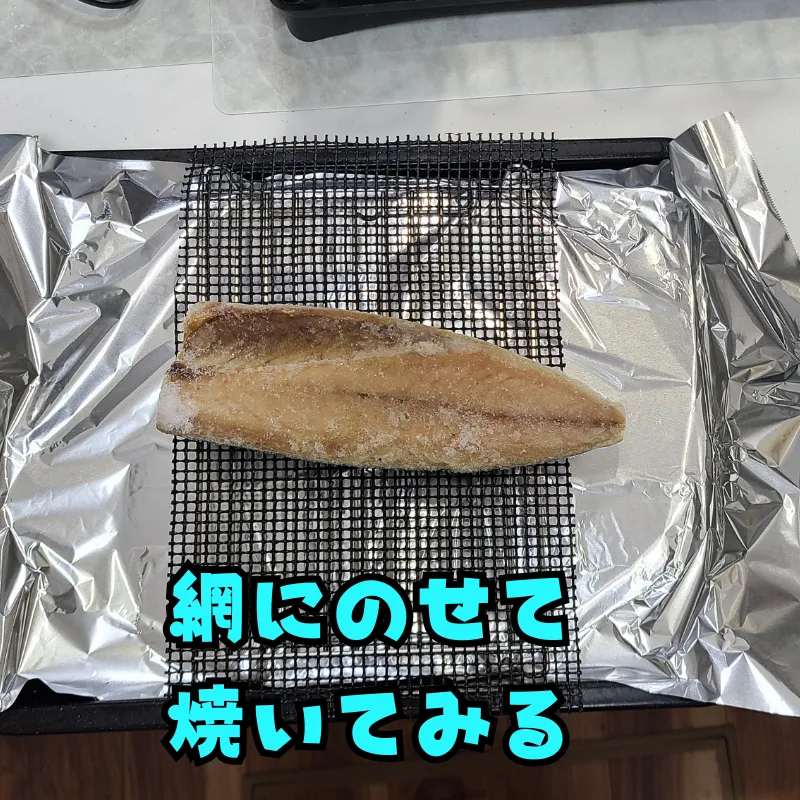 アルミホイルを敷いたトレイの上に、黒いメッシュ状のGWHOLEグリルマットが敷かれ、その上に凍ったサバの半身が置かれている。