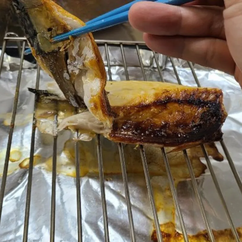 金属製の網にくっついた焼きサバを箸で持ち上げようとしており、皮が剥がれて身が崩れてしまっている。網の下のアルミホイルには焦げた脂や汚れが付着している。