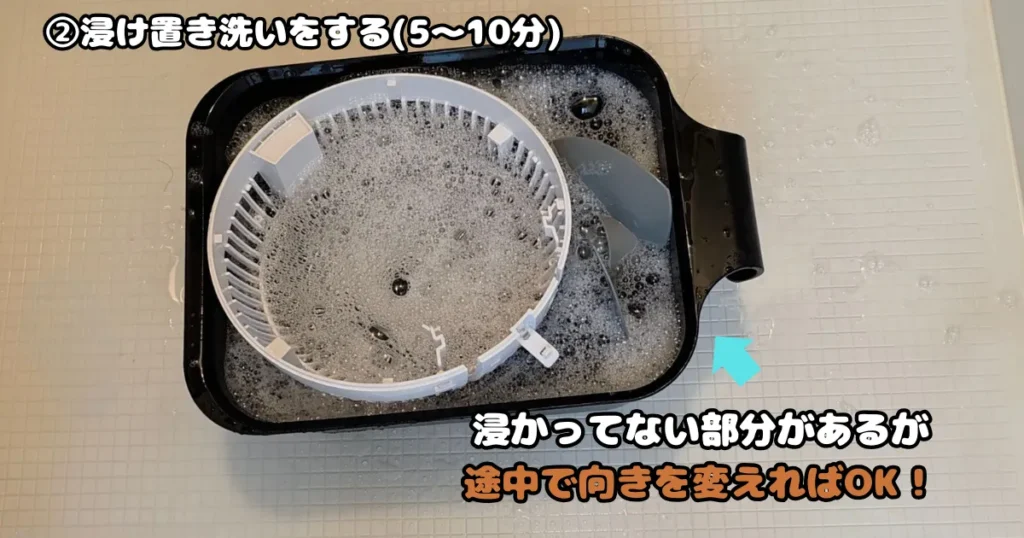 中性洗剤を溶かしたお湯の入った黒い桶に、サーキュレーターの白いカバーやプロペラなどのパーツを入れて浸け置き洗いをしている様子。大きなカバーの一部はお湯からはみ出している。