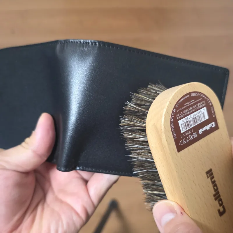 クリームを塗る前に、馬毛ブラシを使って黒い革財布の表面や縫い目のホコリをサッと払い落としている様子。