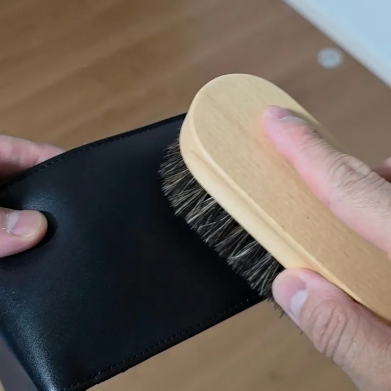 黒い革財布を手に持ち、表面や縫い目の溝の汚れを馬毛ブラシで払い落としている。
