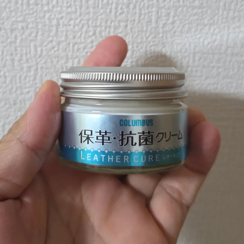 手で持たれた、コロンブスのレザーキュア保革・抗菌クリームの瓶。日常ケアにおいて保湿と除菌が同時に完了するおすすめのアイテム。