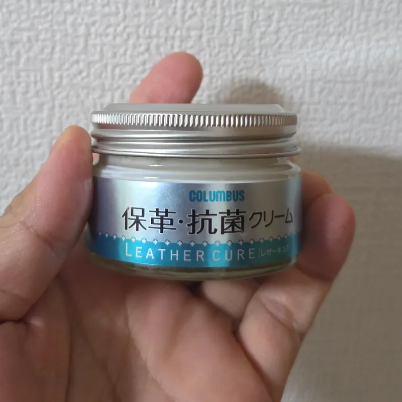 手で持たれた、コロンブスのレザーキュア保革・抗菌クリームの瓶。財布の日常ケアにおいて保湿と除菌が同時に完了するおすすめのアイテム。