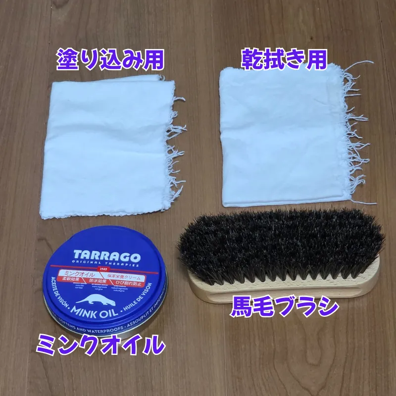 木目調の床の上に並べられた、革財布のお手入れに必要な4つのアイテム。左上に「塗り込み用」と書かれた白い布、右上に「乾拭き用」と書かれた白い布、左下に「ミンクオイル」と書かれた青い缶、右下に「馬毛ブラシ」と書かれたブラシがある。