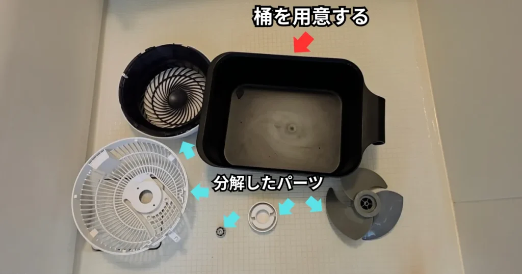 浴室の床に置かれた浸け置き用の黒い桶と、その周りに並べられた前面カバーやプロペラなどの分解済みのサーキュレーターのパーツ。