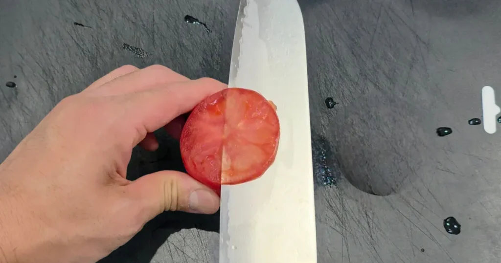 黒いまな板の上で、カットしたトマトを手に持ち、その断面に包丁の刃を添えている様子。