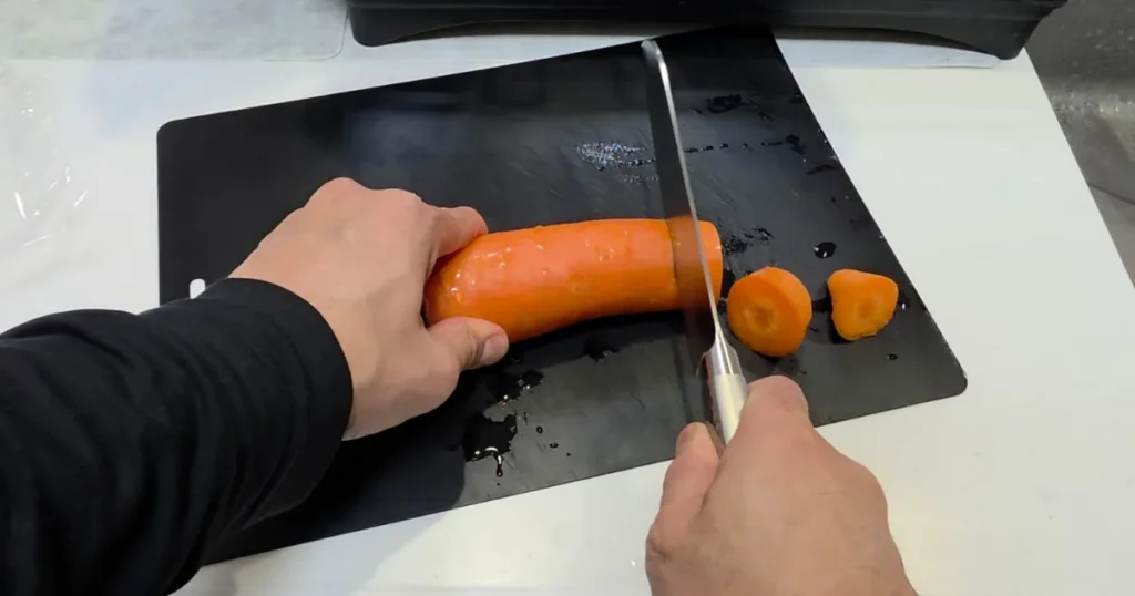黒いまな板の上で、皮を剥いたニンジンを包丁で輪切りにしている様子。