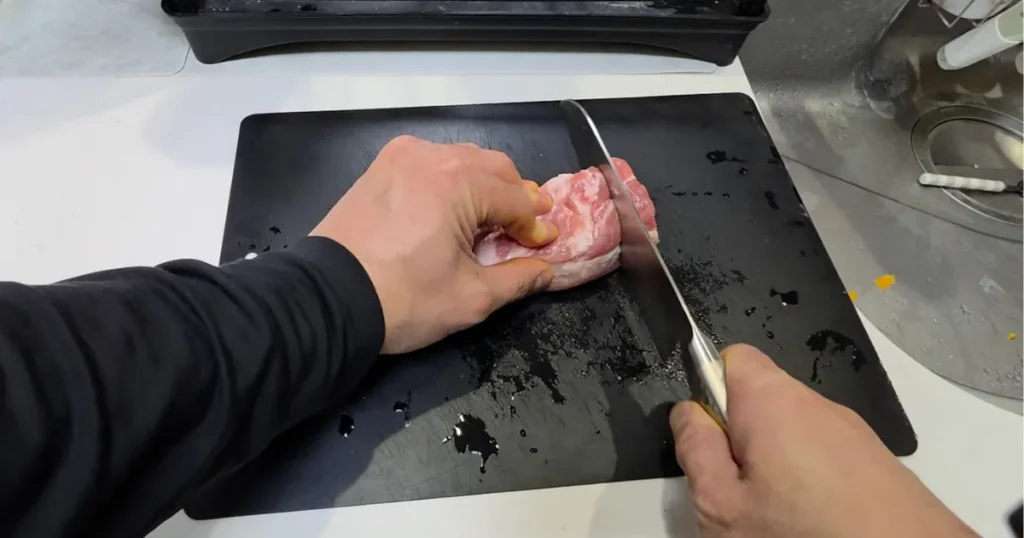 黒いまな板の上で、豚肉の塊を包丁で切っている様子。