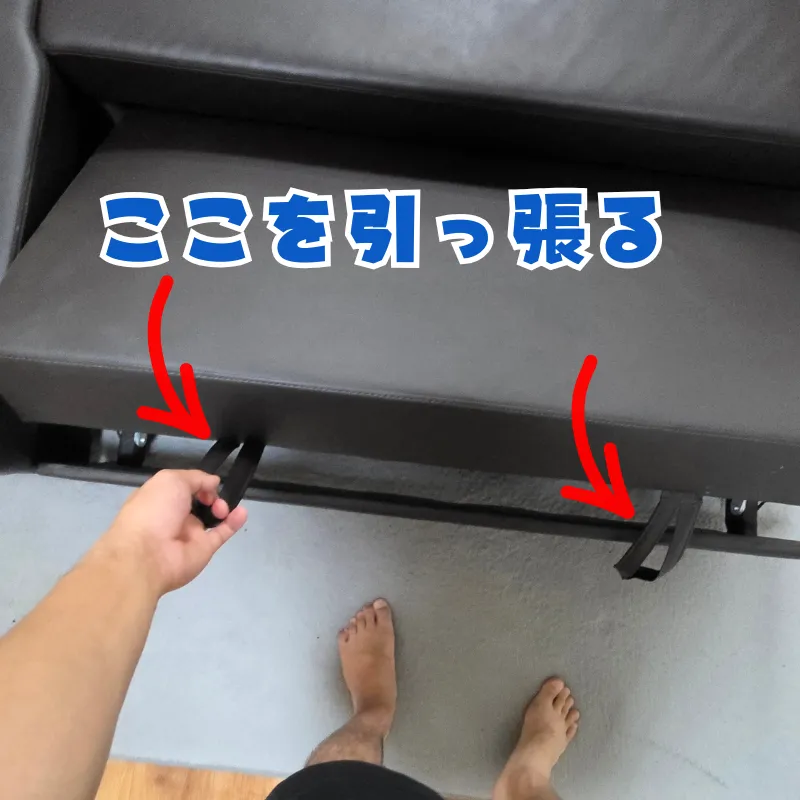 黒いソファの座面下にある取っ手を手前に引っ張って、ベッド部分を引き出している様子。