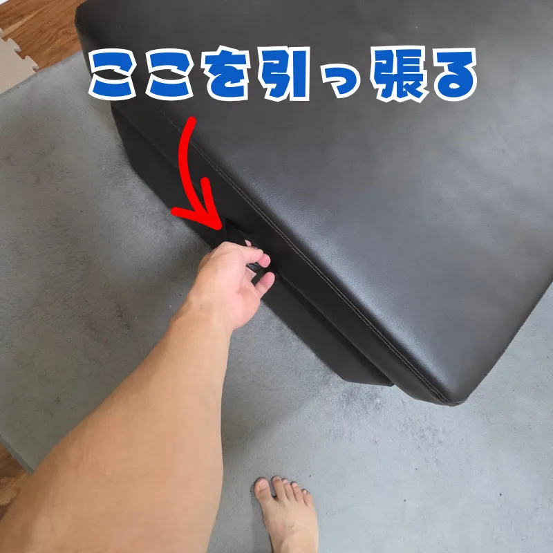 黒いカウチソファの座面下にある取っ手を片手で上に引っ張っている様子。