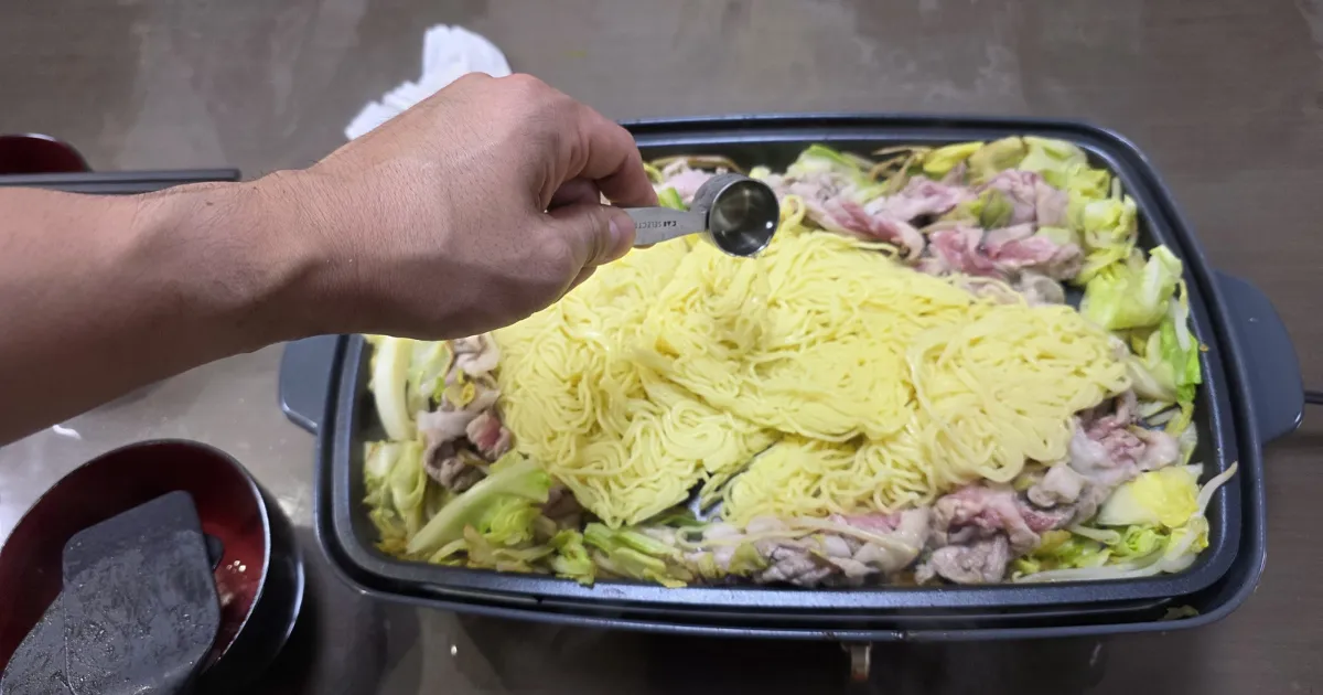ホットプレートで焼きそばを調理している様子。具材の上に計量スプーンで透明な液体を注いでいる。