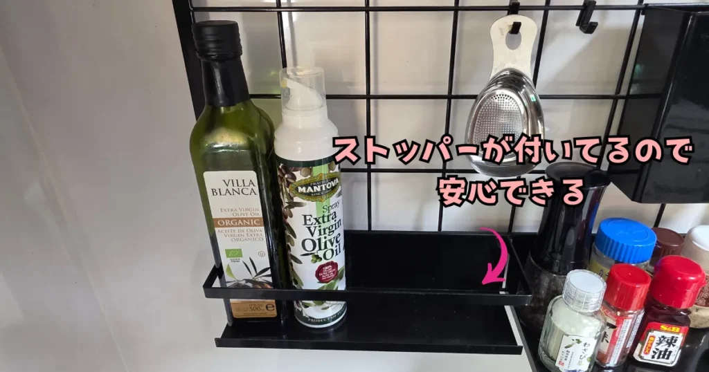 山崎実業(tower)のレンジフードメッシュパネルに取り付けられた、ストッパー付きの黒いラック。オリーブオイルの瓶が2本並んでおり、安定して収納されている。