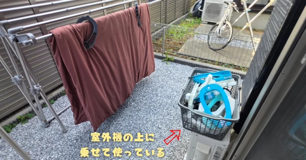 庭やベランダのエアコン室外機の上に、布団バサミを入れた買い物カゴを置いて、しゃがまずに作業する便利なアイデア。