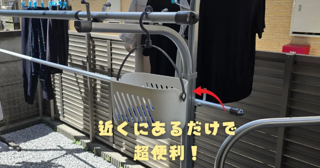 洗濯物が干された物干し竿に、大きなフックで直接掛けられた、洗濯バサミ入りの白いプラスチック製バスケット。