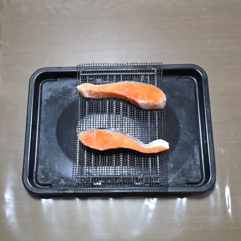 黒い角皿の上に網と黒いグリルマットを敷き、凍った鮭の切り身を2切れ並べている状態。